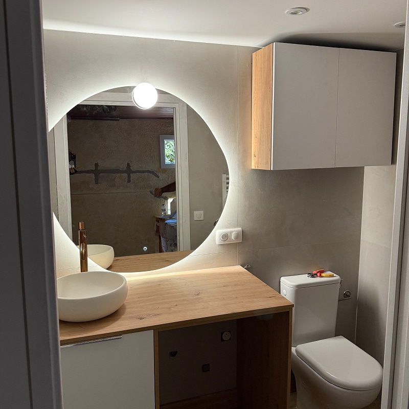 Salle d'eau moderne : miroir rond lumineux, vasque blanche sur plan de travail en bois clair, robinet laiton et meuble mural.