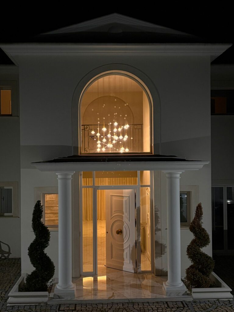 Entrée de Maison de Luxe : Lustre Cristal et Colonnes Entrée de maison luxueuse avec lustre en cristal, colonnes blanches et arbustes sculptés éclairés la nuit.