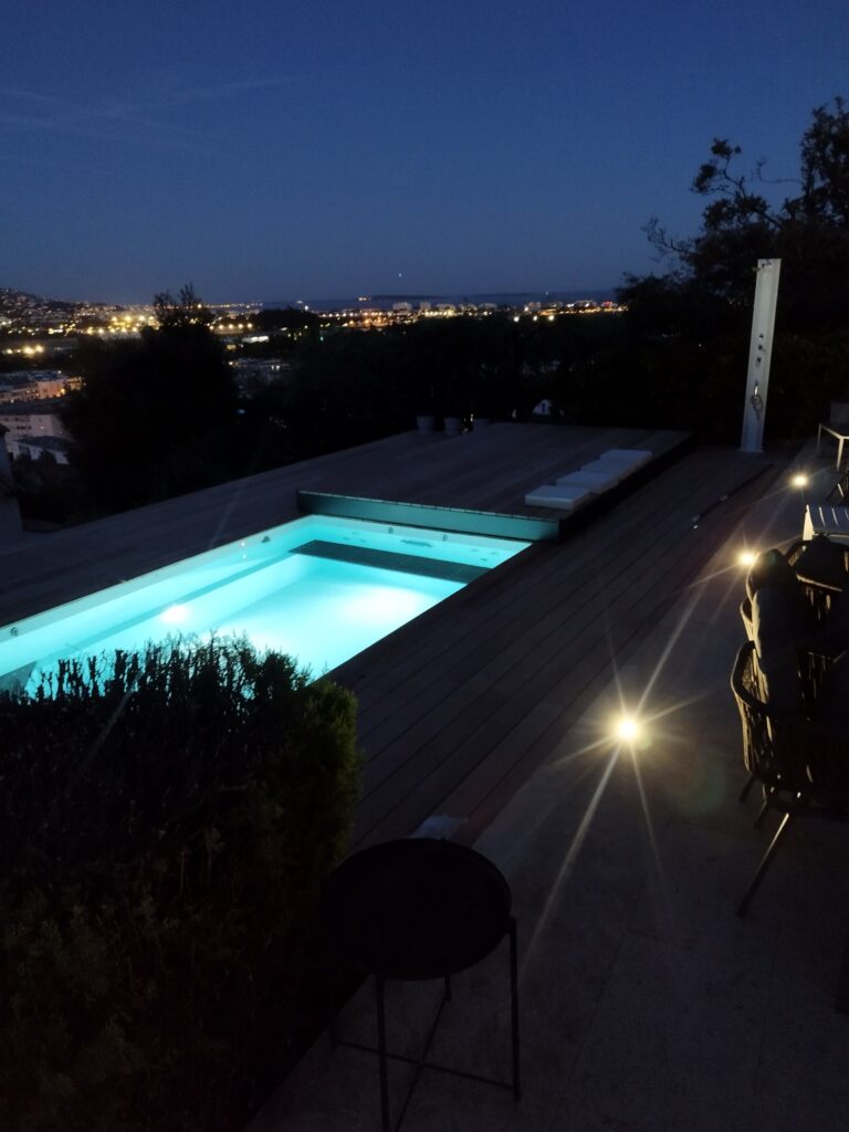 Piscine éclairée sur une terrasse en bois éclaire a nuit avec vue panoramique sur les lumières de la ville.