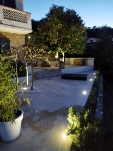 Terrasse en pierre éclairée de nuit avec mobilier, grands arbres et plantes en pot dans un jardin moderne.